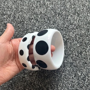 May include: Two thick, circular bangle bracelets made of smooth white material, each featuring a pattern of large, solid black polka dots distributed across the exterior surface. The bracelets have a glossy finish and a wide, rounded profile.
