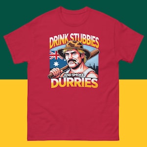 May include: Red t-shirt with a cartoon illustration of a man wearing a hat and holding a baseball bat. The text "Drink Stubbies and Smoke Durries" is printed on the shirt.