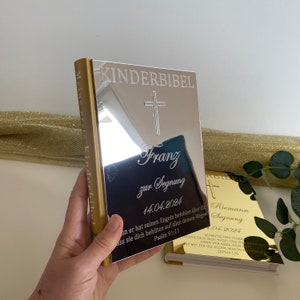 May include: A silver-colored book with the words "Kinderbibel" and a cross engraved on the front cover. The book is personalized with the name "Franz" and the date "14.04.2024" for a christening or baptism.