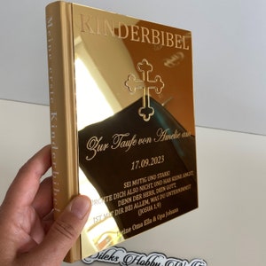 May include: A gold-colored book with the title "Kinderbibel" and the text "Meine erste Kinderbibel" on the spine. The front cover features a cross and the text "Zur Taufe von Amelie am 17.09.2023". The text "Sei mutig und stark!" is also visible on the cover.