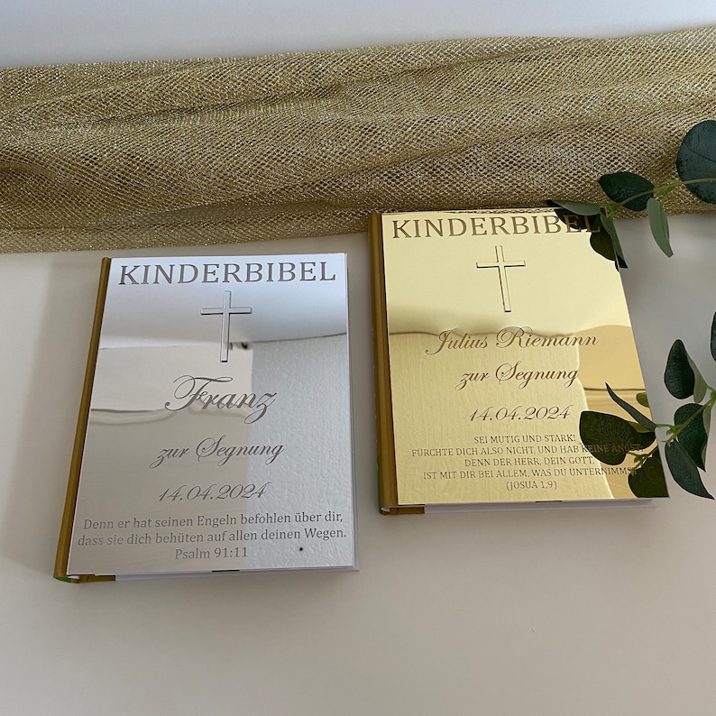 May include: Two silver and gold children's Bibles with personalized inscriptions. The silver Bible has the name "Franz" and the date "14.04.2024" inscribed on it. The gold Bible has the name "Julius Riemann" and the date "14.04.2024" inscribed on it. Both Bibles have a cross and a Bible verse inscribed on them.