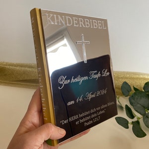 May include: A silver book with the words "KINDERBIBEL" and "Meine erste Kinderbibel" engraved on the cover. The book is inscribed with the words "Zur heiligen Taufe Leon am 14. April 2024" and a Bible verse: "Der HERR beh&uuml;tet dich vor allem B&ouml;sen, er beh&uuml;tet dein Leben." Psalm 121,7
