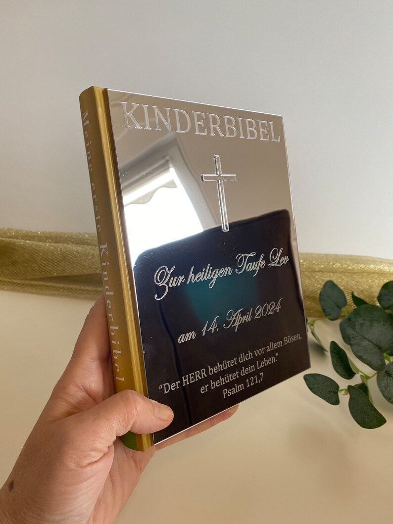 May include: A silver book with the words "KINDERBIBEL" and "Meine erste Kinderbibel" engraved on the cover. The book is inscribed with the words "Zur heiligen Taufe Leon am 14. April 2024" and a Bible verse: "Der HERR beh&uuml;tet dich vor allem B&ouml;sen, er beh&uuml;tet dein Leben." Psalm 121,7