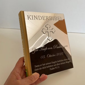May include: A gold-colored book with the title "KINDERBIBEL" engraved on the front cover. The book is personalized with the text "Zur Taufe von Paulina am 03. Oktober 2023" and a quote from Psalm 91:11.