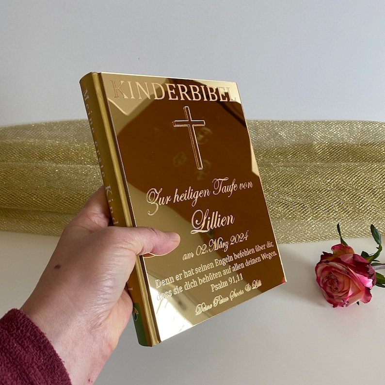May include: A gold-colored book with the words "KINDERBIBER" and "Zur heiligen Taufe von Lillien" engraved on the cover. The book also has a cross and a quote from Psalm 91:11.