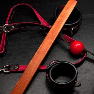 Puede incluir: Una paleta de madera con una correa de cuero roja y puños de cuero negro. La paleta está hecha de madera natural y tiene un acabado liso. La correa y los puños son ajustables y tienen hebillas metálicas.