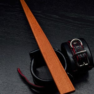 Puede incluir: Una paleta de madera con un acabado liso de color marrón rojizo, que se encuentra sobre una superficie negra. También hay dos correas de cuero negro con costuras rojas y hebillas plateadas sobre la superficie.