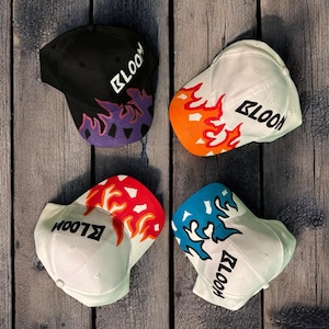 Może przedstawiać: Cztery białe czapki bejsbolowe z różnokolorowymi płomieniami z tyłu. Każda czapka ma napis "BLOOM" w kolorze czarnym z przodu.