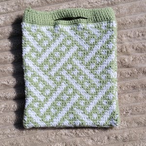 May include: A handmade crochet bag in a geometric pattern of white and light green. The bag has a solid light green border and a small handle at the top. The bag is sitting on a textured, tan surface.