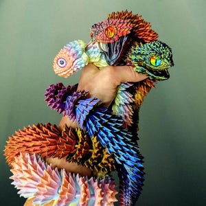Puede incluir: Una mano sostiene tres esculturas de serpientes articuladas y coloridas. Las serpientes son en tonos naranja, rojo, azul, morado y verde. Cada serpiente tiene una cabeza detallada con mandíbulas abiertas y ojos vibrantes. Las escamas son texturizadas y superpuestas.