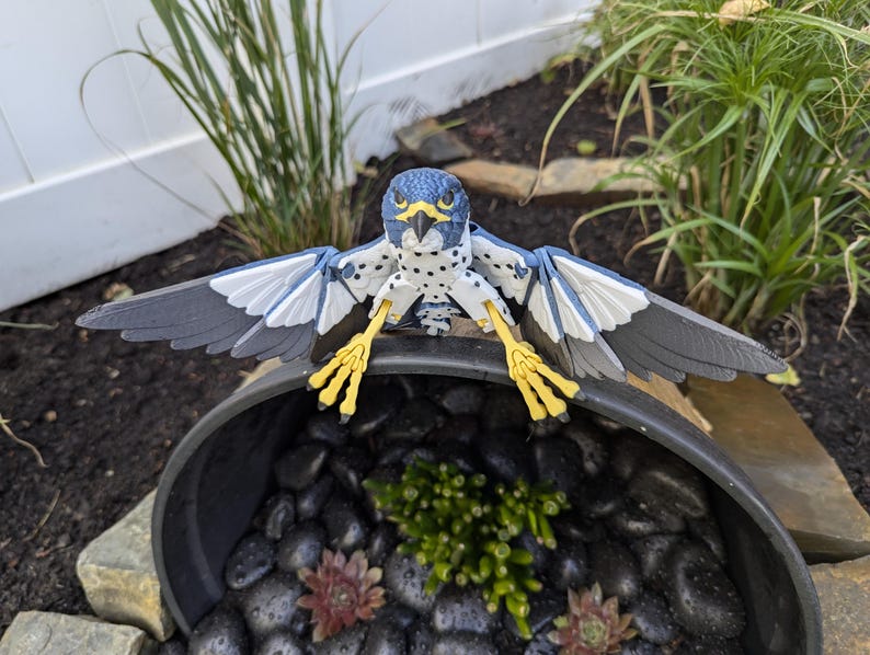 May include: A blue and white toy bird with yellow feet perched on the edge of a black planter. The planter is filled with small black stones and green plants.