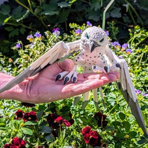 May include: A white and grey bird figurine with black eyes and talons. The bird is perched on a hand with its wings spread out.