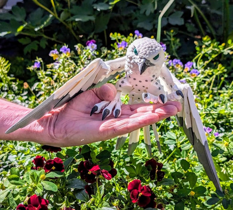 May include: A white and grey bird figurine with black eyes and talons. The bird is perched on a hand with its wings spread out.