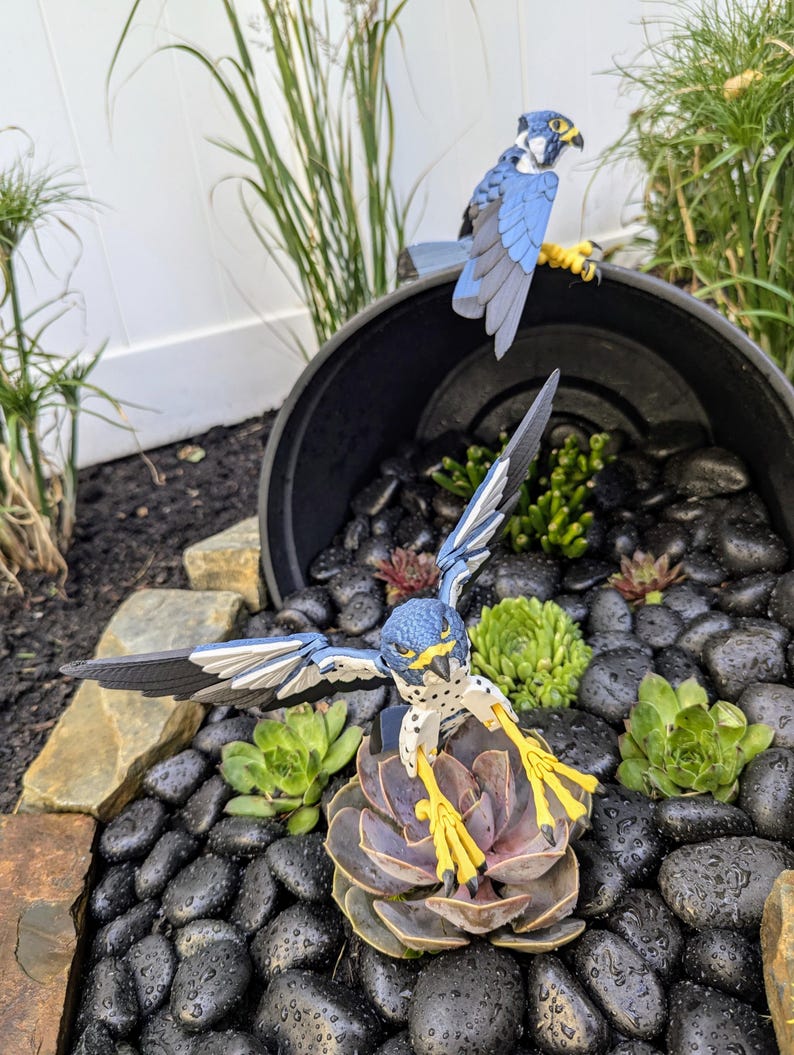 May include: Two blue and grey bird figurines perched on a rock garden. The bird on the left is perched on a succulent plant with its wings spread. The bird on the right is perched on a black pot.