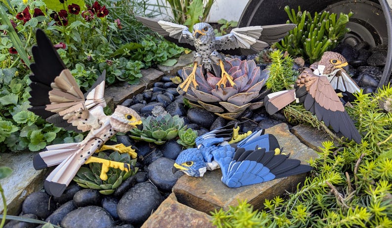 May include: Four papercraft birds in a garden setting. The birds are a brown and white kestrel, a blue and black bird, a brown and white kestrel, and a brown and white bird with a black head.