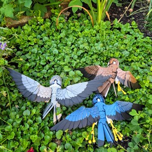 May include: Three papercraft birds with outstretched wings, in shades of grey, blue, and brown, are displayed on a bed of green foliage. The birds are intricately detailed and appear to be crafted from paper.