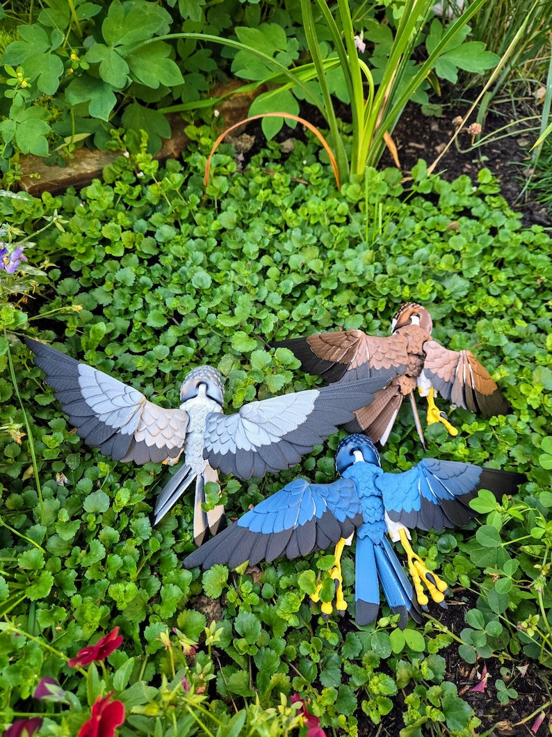 May include: Three papercraft birds with outstretched wings, in shades of grey, blue, and brown, are displayed on a bed of green foliage. The birds are intricately detailed and appear to be crafted from paper.