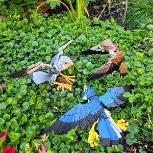 May include: Three papercraft birds with outstretched wings, a grey and white bird, a brown bird, and a blue bird, are posed on a bed of green clover.