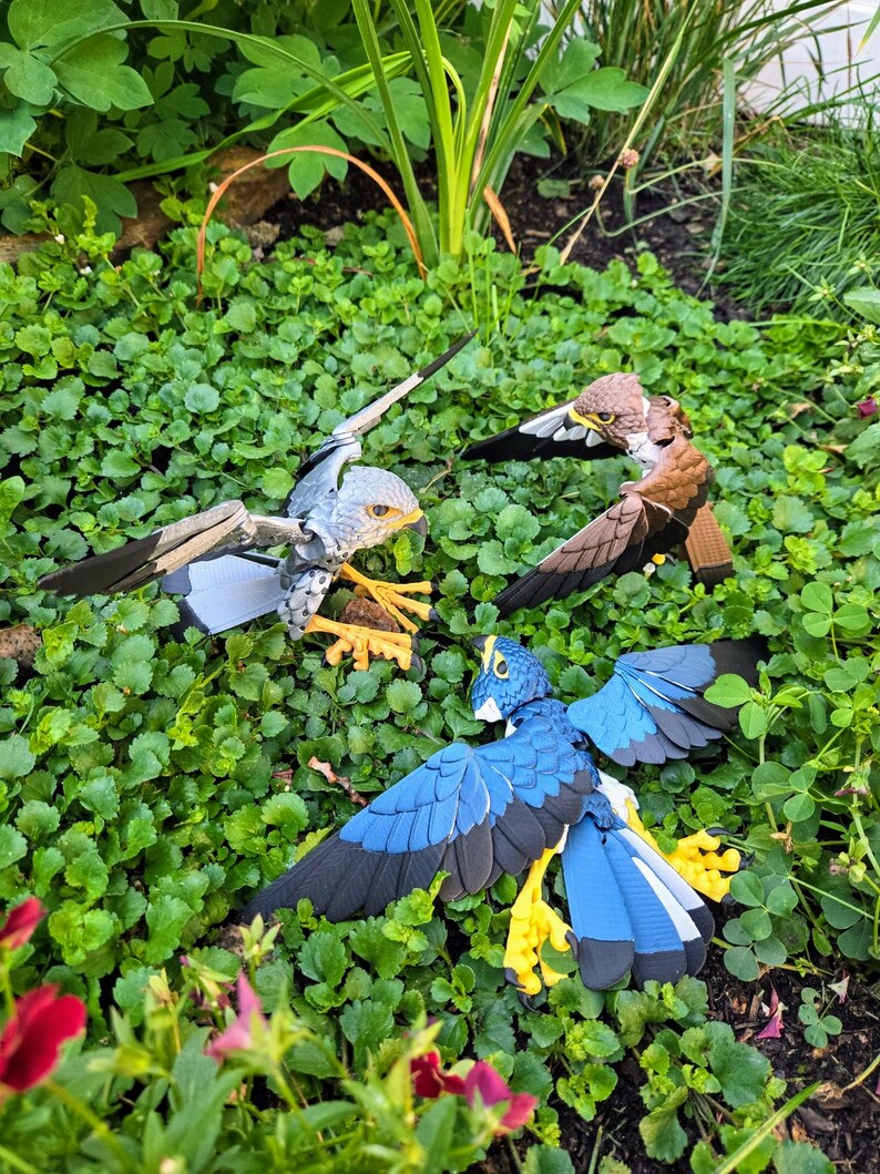 May include: Three papercraft birds with outstretched wings, a grey and white bird, a brown bird, and a blue bird, are posed on a bed of green clover.