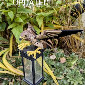 May include: A brown and black bird figurine perched on a black metal lamp post with a clear glass enclosure. The bird has yellow feet and black spots on its body. The background is a green leafy garden.