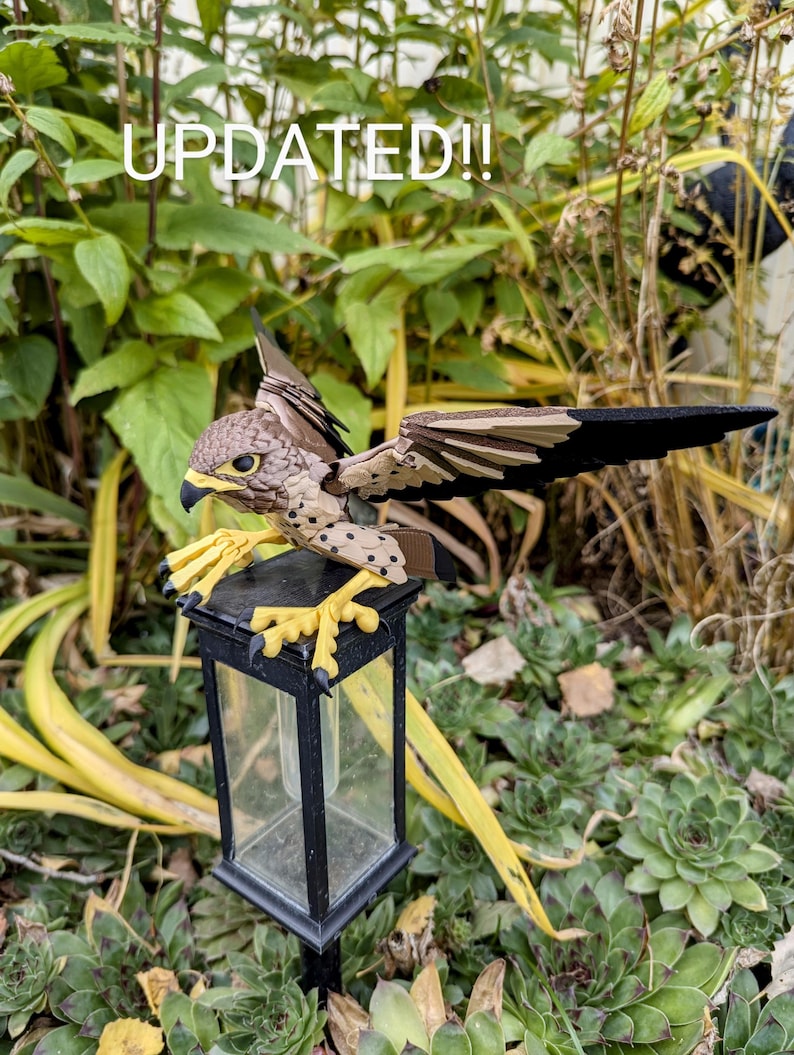 May include: A brown and black bird figurine perched on a black metal lamp post with a clear glass enclosure. The bird has yellow feet and black spots on its body. The background is a green leafy garden.