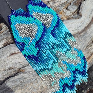 May include: Handmade beaded earrings in shades of blue and silver. The earrings feature a fringe design with a heart-shaped pattern in the center. The earrings are displayed on a black card that says "handmade with love".