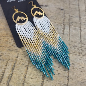 May include: Handmade beaded earrings with a mountain design. The earrings feature gold-colored circular pendants with a mountain silhouette. The fringe is white, gold, and teal, creating a layered effect. The earrings hang from gold-colored ear wires.