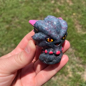 May include: A detailed sculpture of a creature's head, featuring a dark gray and purple speckled body with pink accents on the horns and red accents on the lower portion. The creature has yellow eyes with red pupils. The sculpture is held in a hand.