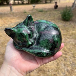 May include: A polished stone carving of a sleeping cat, crafted from dark green zoisite with prominent inclusions of deep magenta ruby. The sculpture features smooth, rounded contours and a glossy finish, depicting the feline curled in a resting pose.