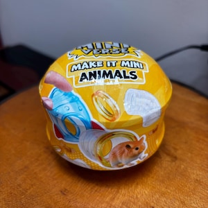 Puede incluir: Un recipiente amarillo y redondo con el texto "MAKE IT MINI ANIMALS". Contiene imágenes de un hámster, una estructura azul y blanca y anillos dorados. El producto es probablemente un juguete o un kit de manualidades.
