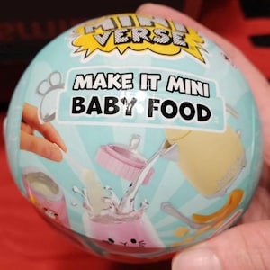 Pode incluir: Uma bola Mini Verse azul clara com o texto "MAKE IT MINI BABY FOOD". A bola apresenta ilustrações de biberões, um aquecedor de biberões e comida para bebés. A parte superior da bola tem as palavras "MINI VERSE" numa explosão amarela.