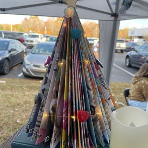 May include: A handmade Christmas tree made from folded magazine pages, adorned with heart-shaped ornaments and string lights. The tree is topped with a gold star and sits on a blue book. A white candle is in the foreground.