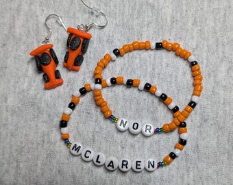 May include: Two orange and black car charm earrings and a beaded bracelet with the words "McLaren" and "Nor" in white letters.