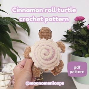 Può includere: Una tartaruga all'uncinetto con un design a girella alla cannella. La tartaruga ha una testa e arti marroni, e un guscio con una spirale color crema e un centro lilla chiaro. Il testo "Cinnamon roll turtle crochet pattern" è in alto, e "pdf pattern" è in un riquadro viola.