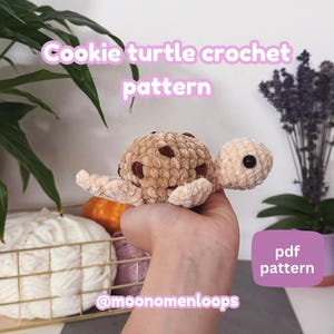 May include: A crocheted turtle with a cookie-like shell, featuring brown chocolate chip details. The turtle's head and limbs are a light beige color. Text on the image reads "Cookie turtle crochet pattern" and "pdf pattern". The Etsy shop handle @moonomenloops is also visible.