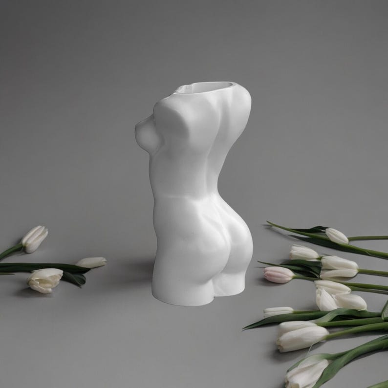 May include: White ceramic vase shaped like a human torso. The vase is empty and has a smooth, matte finish. The vase is sitting on a gray surface with white tulips scattered around it.