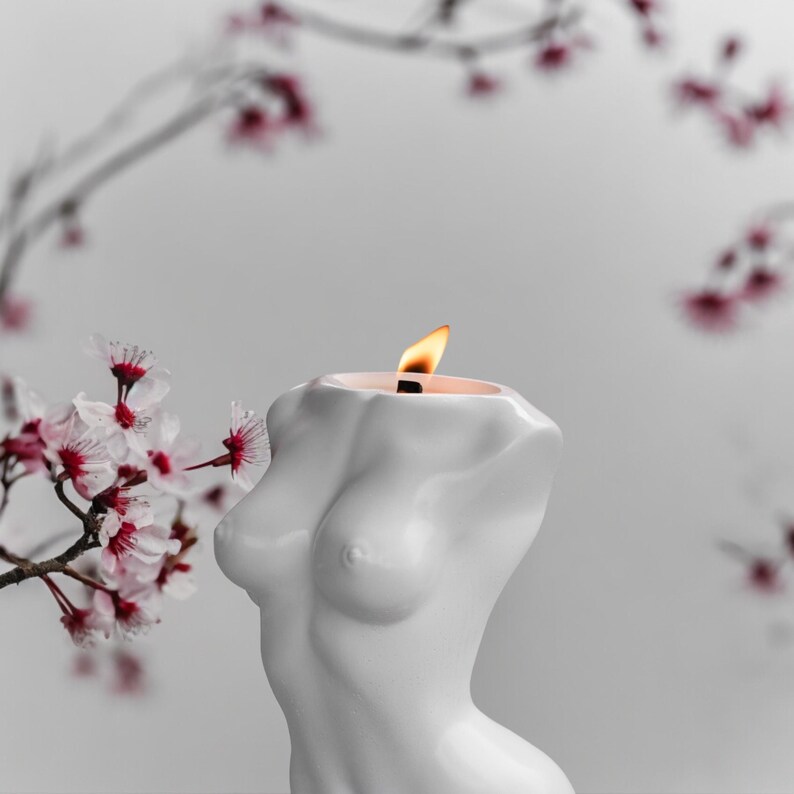 May include: A white candle shaped like a human torso with a lit flame on top. The candle is in front of a white background with a blurred image of pink flowers.