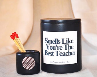 May include: A black candle and match holder set. The candle has a white label with the text "Smells Like You're The Best Teacher". The match holder has a circular design with a pattern of maroon dots. The candle is 8oz.