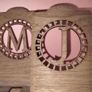 Op de afbeelding: Twee houten telefoonstandaards met een decoratief cirkelontwerp en de letters "M" en "J" uitgesneden. De standaards zijn gemaakt van lichtbruin hout en hebben een rustieke afwerking.