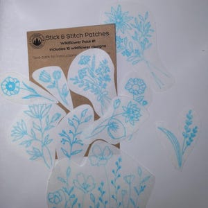 Puede incluir: Un paquete de 10 parches de bordado de flores silvestres azules sobre tela blanca. Los parches están etiquetados con "Stick & Stitch Patches Wildflower Pack #1 Includes 10 wildflower designs" y "see back for instructions".