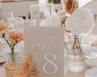 May include: A beige table number card with the text "Table 8" in white script font. The card is on a clear acrylic stand and is surrounded by white and beige flowers in glass vases.