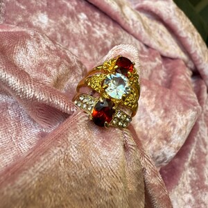 May include: A gold-toned ring featuring a large central clear round stone flanked by two oval-shaped deep red stones. The band is embellished with small clear crystals and intricate metalwork, creating a layered appearance with multiple bands.