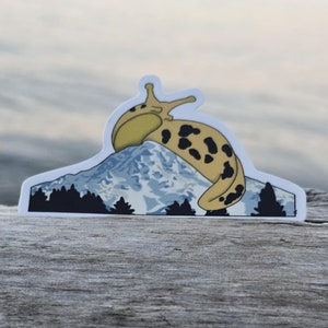 May include: A die-cut sticker featuring a yellow and black spotted banana slug atop a mountain range with a blue and white color scheme. The sticker has a white border and is placed on a weathered wooden surface.