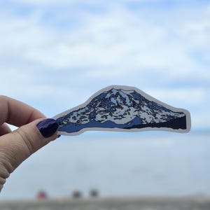 May include: A blue and white sticker of a mountain with snow on the top. The sticker is being held in a hand against a blue sky background.