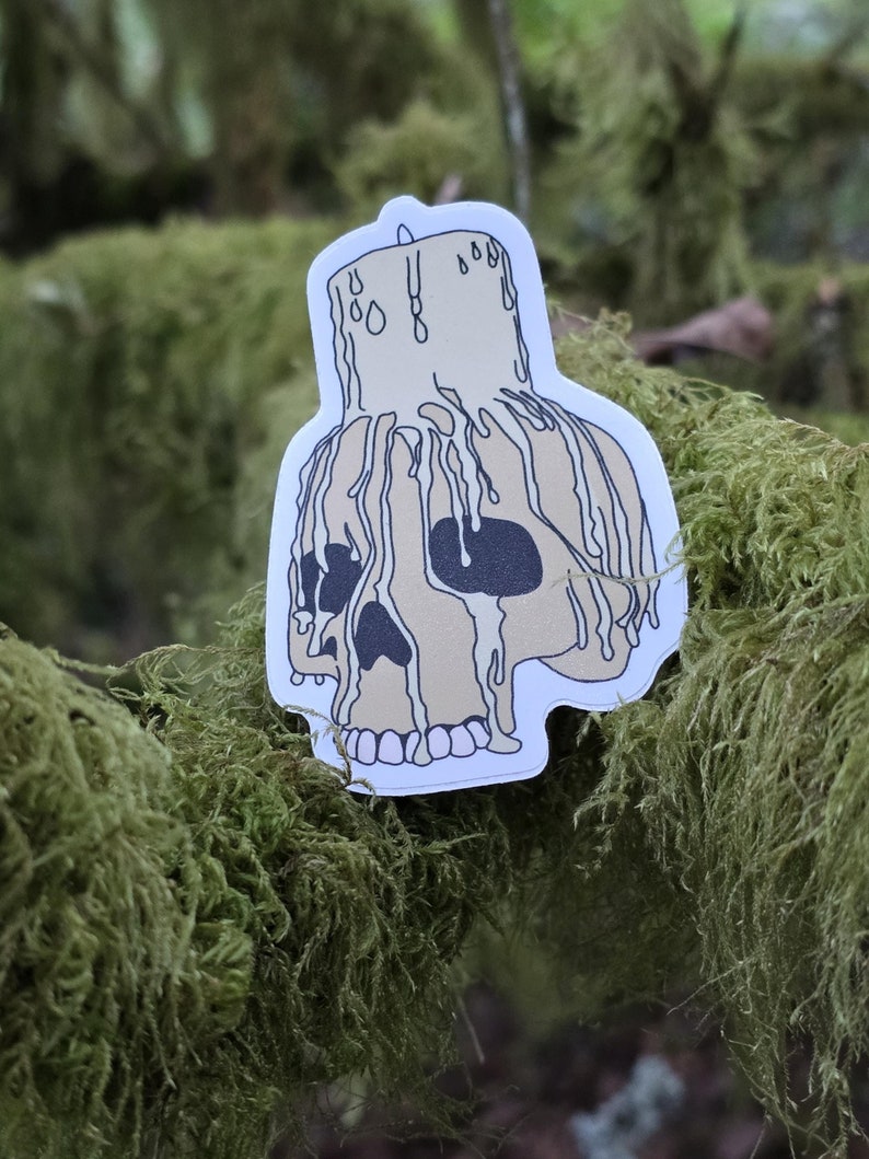 May include: A white sticker with a black outline of a skull with a candle on top. The candle is dripping wax onto the skull. The sticker is on a green mossy surface.