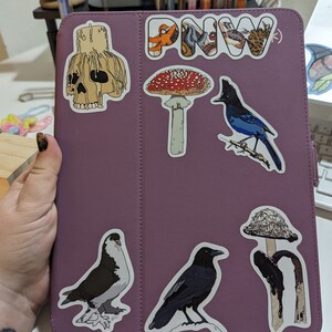May include: A purple iPad case with various stickers, including a skull, a mushroom, a bird, a raven, a pigeon, and a mushroom with a black stem. The case also has a sticker that says "PNW".