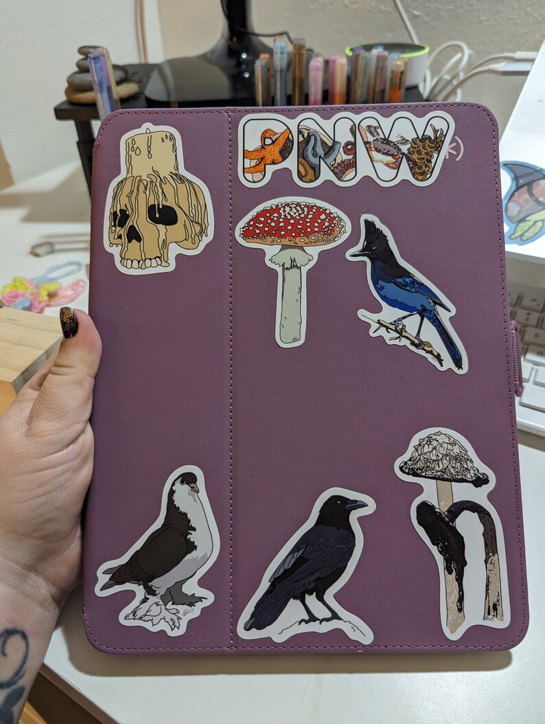 May include: A purple iPad case with various stickers, including a skull, a mushroom, a bird, a raven, a pigeon, and a mushroom with a black stem. The case also has a sticker that says "PNW".