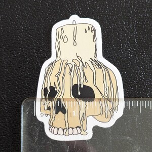 May include: A white skull with dripping wax, resembling a candle, with black eye sockets and teeth. The wax is dripping down the skull's face and onto the base.