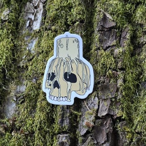 May include: A white sticker with a cartoon illustration of a skull with a dripping candle on top. The skull is yellow with black eyes and a black outline. The candle is yellow with black wax dripping down the sides.