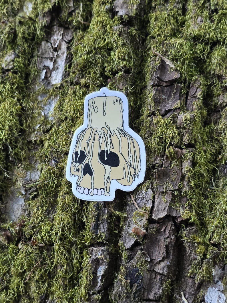 May include: A white sticker with a cartoon illustration of a skull with a dripping candle on top. The skull is yellow with black eyes and a black outline. The candle is yellow with black wax dripping down the sides.